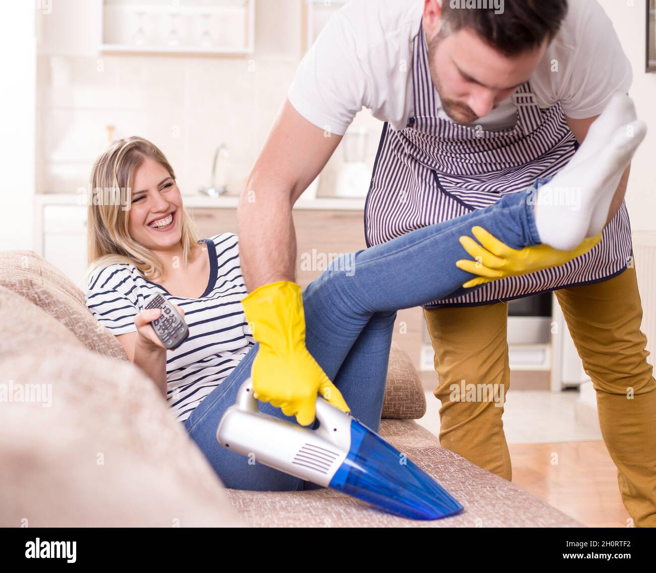 Young man with apron vacuuming sofa with attractive woman sitting and watching tv. Woman enjoying while husband doing chores Stock Photo