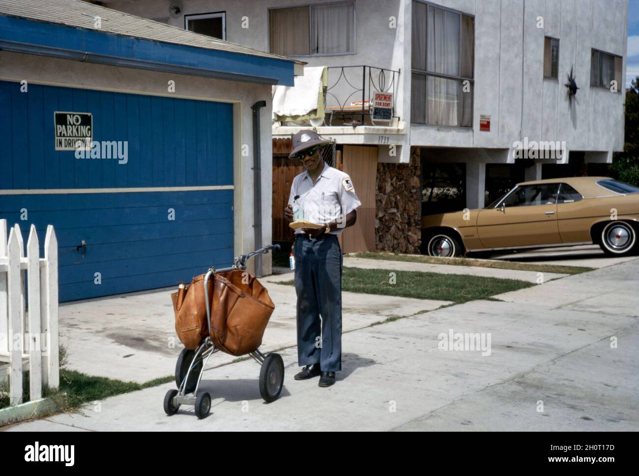 An Afro-American postman delivering the US mail in suburban Long Beach, California, USA in 1972. He has with him a stylish, leather 4-wheeled, push hand-cart or trolley. A wide-brimmed, sun protection hat is part of the uniform. A mail carrier, mailman, mailwoman, postal carrier, postman, postwoman, or letter carrier is an employee of a post office or postal service. The United States Postal Service (USPS, the Post Office, US Mail, or Postal Service) is an agency of the federal government. This image is from an old American Kodak amateur colour transparency – a vintage 1970s photograph. Stock Photo