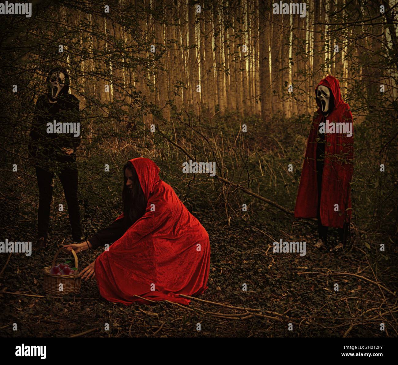 Little red riding Hood with masked person in black standing behind. Shot in shady woodland. Stock Photo