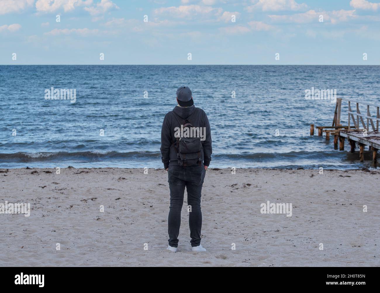 rear view of man wearing hooded sweater and jeans looking at the sea Stock Photo