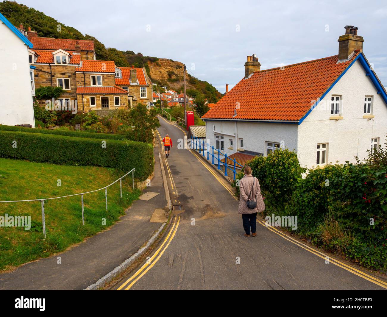 A postman and a senior lady walking into the cliffside village of Runswick Bay with quaint houses and cottages mainly for holiday lets or second homes Stock Photo