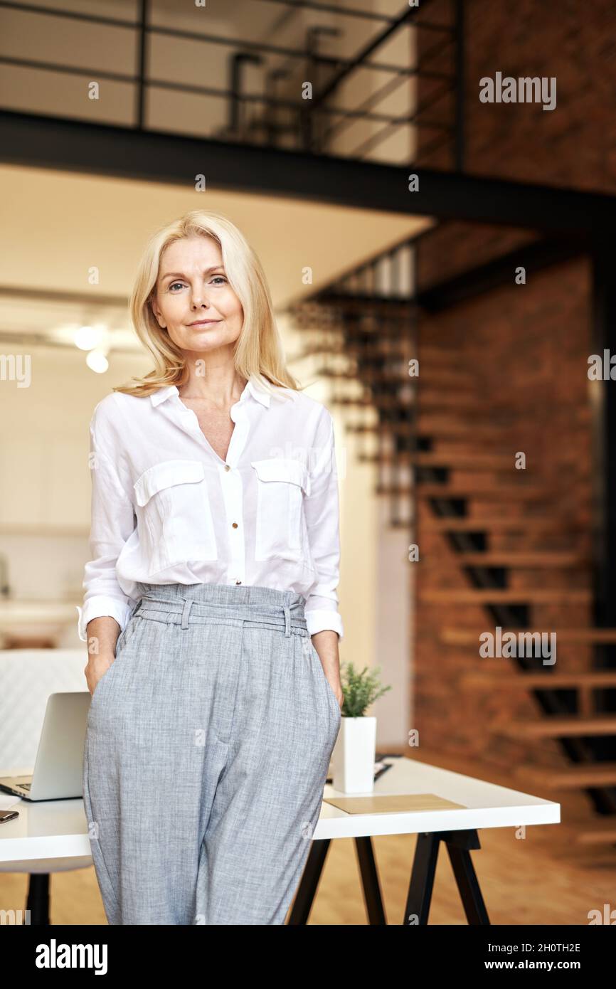 Stylish mature blonde business lady looking away while posing, standing in the office Stock Photo
