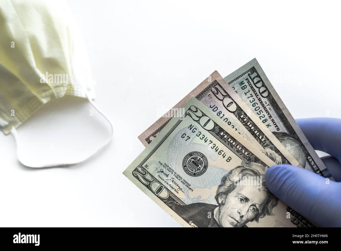 Close up of person wearing a blue latex glove holding a hundred, fifty and twenty dollar bill fanned out and isolated with a yellow face mask or cover Stock Photo