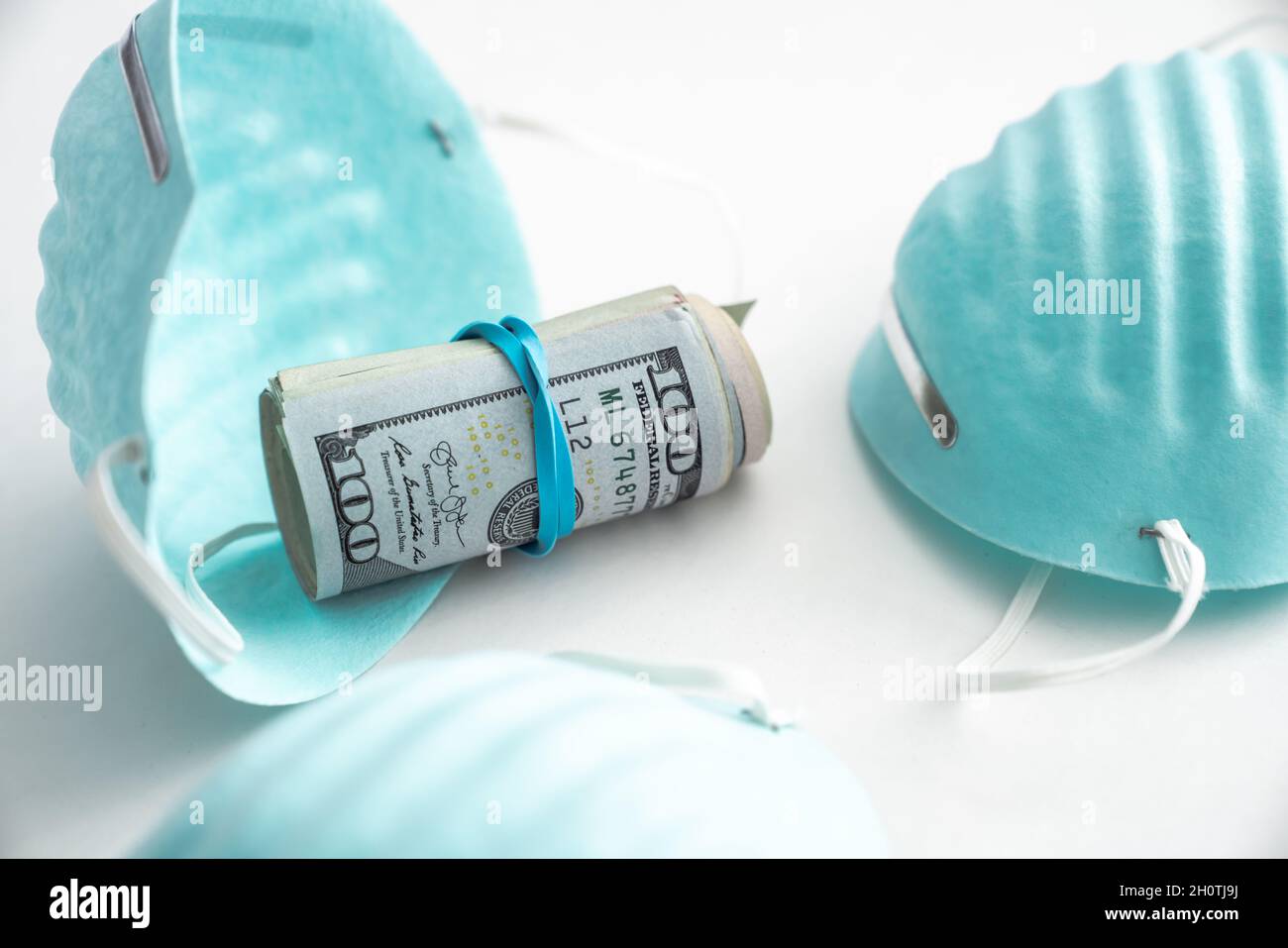 A rolled up wad of cash with a rubberband around it sits in a blue ribbed medical mask or protective face covering used for COVID-19 virus pandemic is Stock Photo