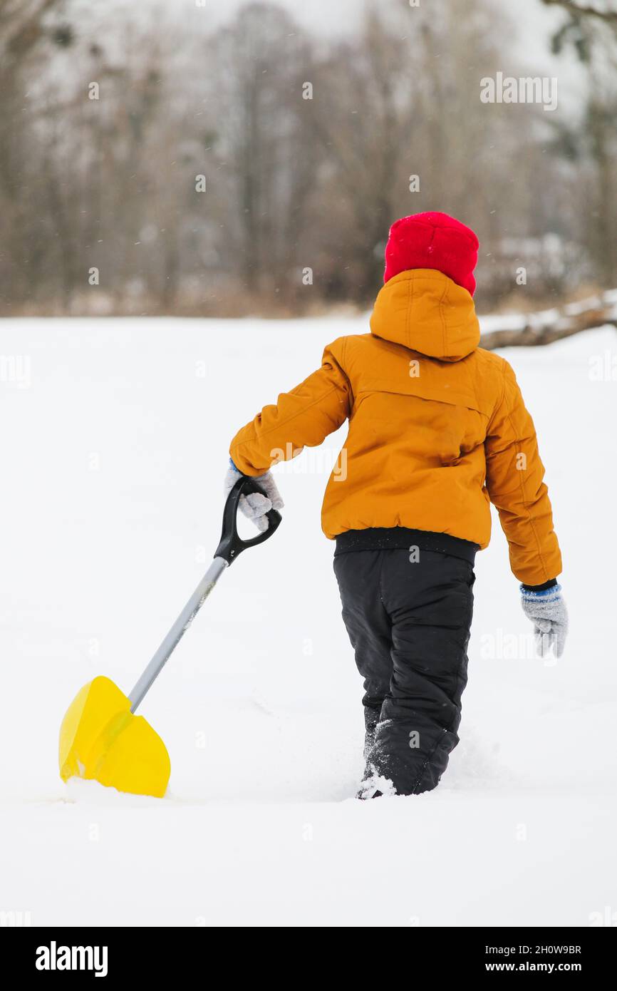 back view of boy in yellow jacket and red hat with scarf with shovel in his hands on a snowy field Stock Photo