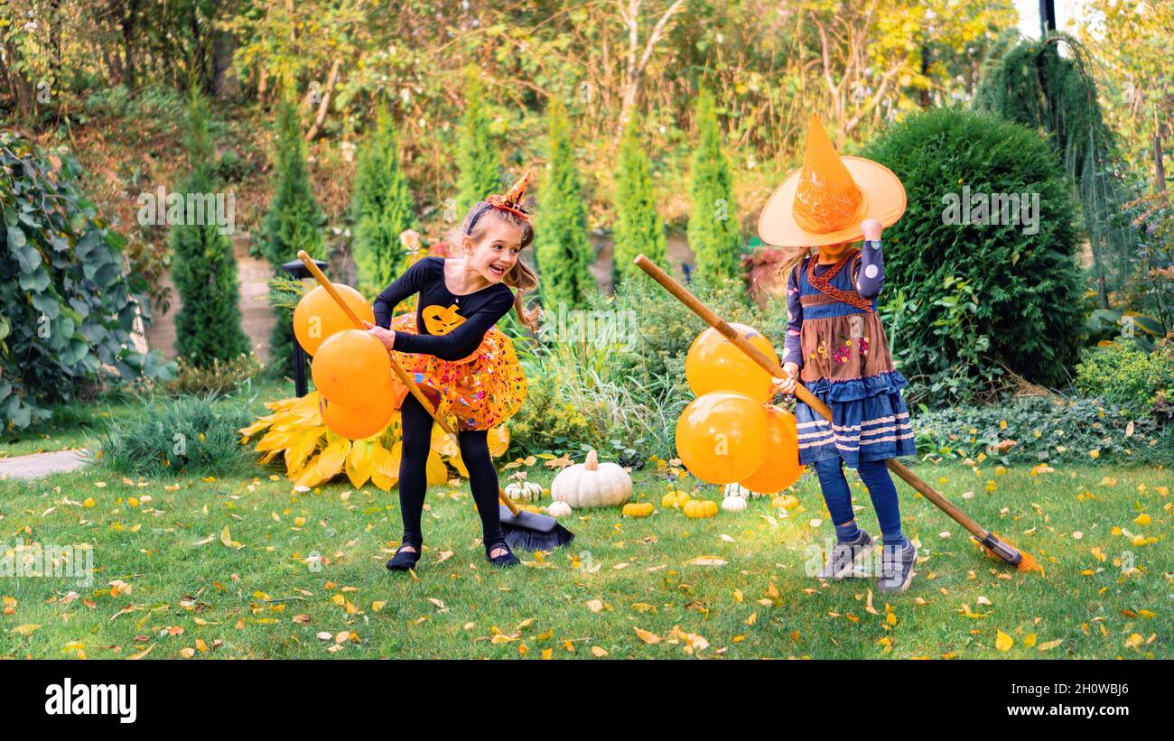 Kids laugh and have fun playing witch with broomsticks for Halloween. Cheerful Halloween party with carnival costumes outdoors. Little witches learn t Stock Photo