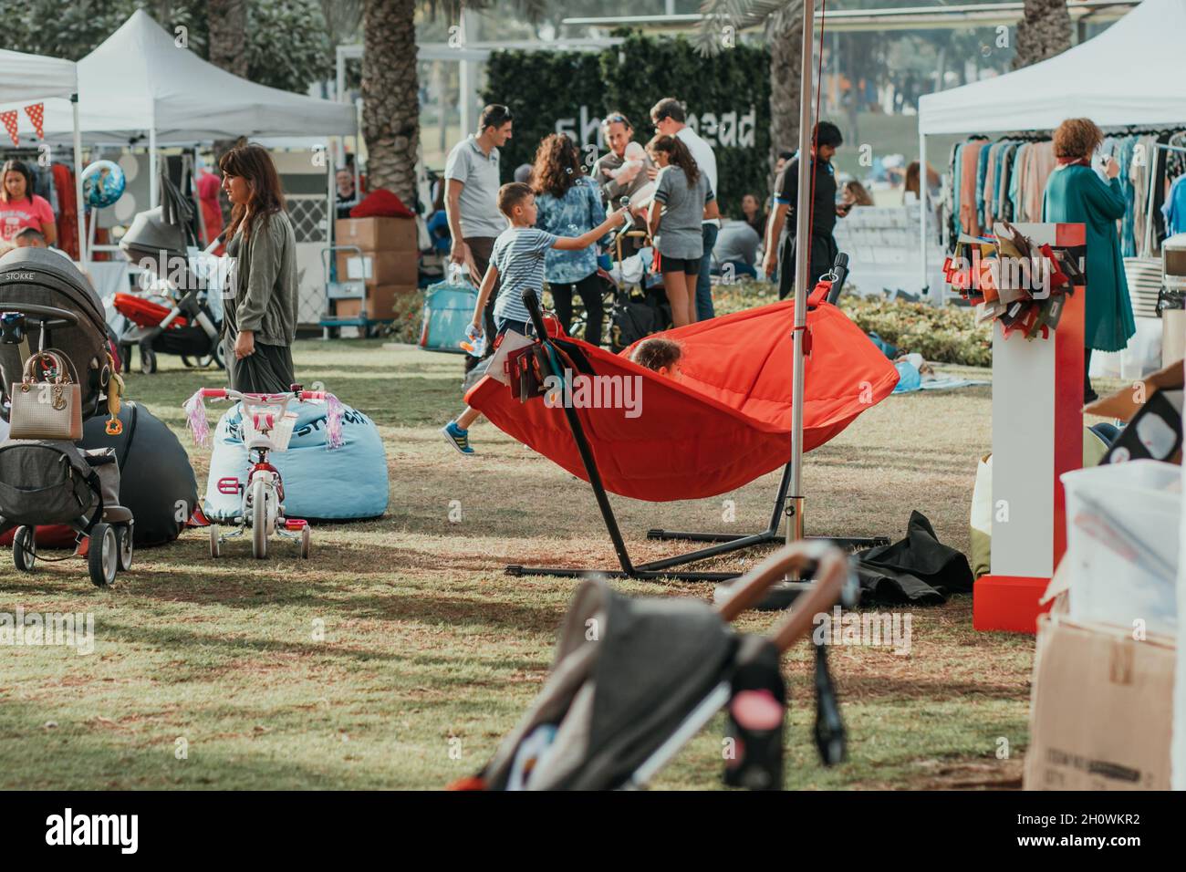 People walking at the park. Kids play and adults go around in the fair with green grass, palms, emerald trees, interesting flag decoration. Stock Photo