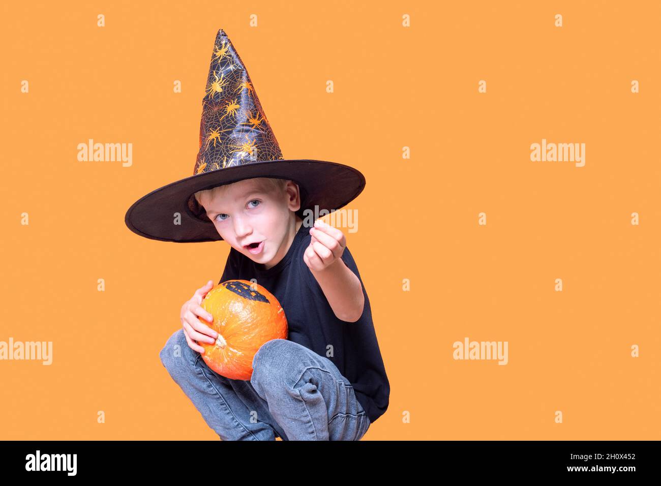Halloween kids. Little Emotional boy in a wizard hat holding an orange pumpkin in his hands and showing a candy on an orange background. The kid wants Stock Photo