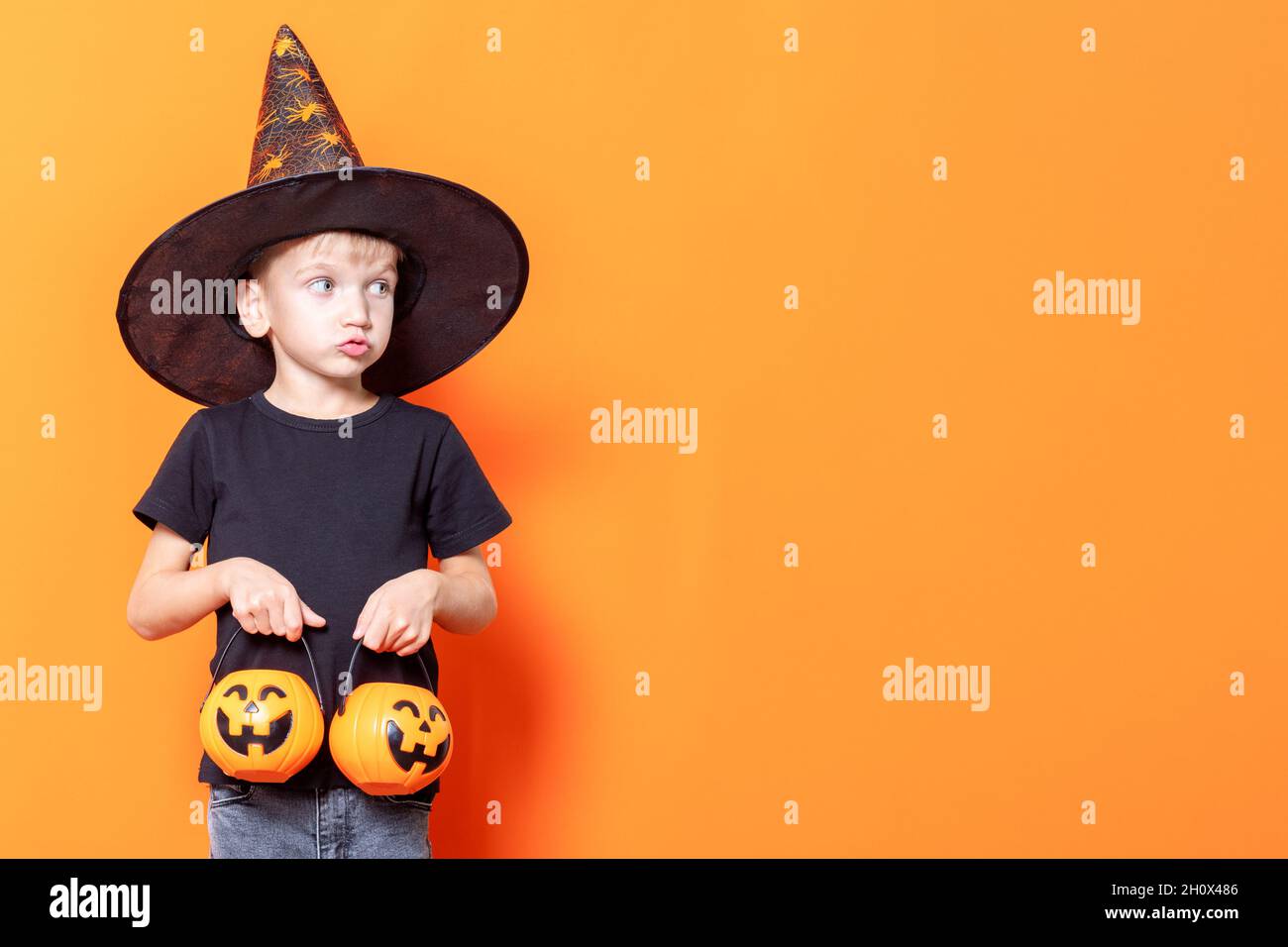 Halloween kids. Little boy in a wizard's hat holding a basket in the shape of a pumpkin with sweets on an orange background, copy space. The kid wants Stock Photo