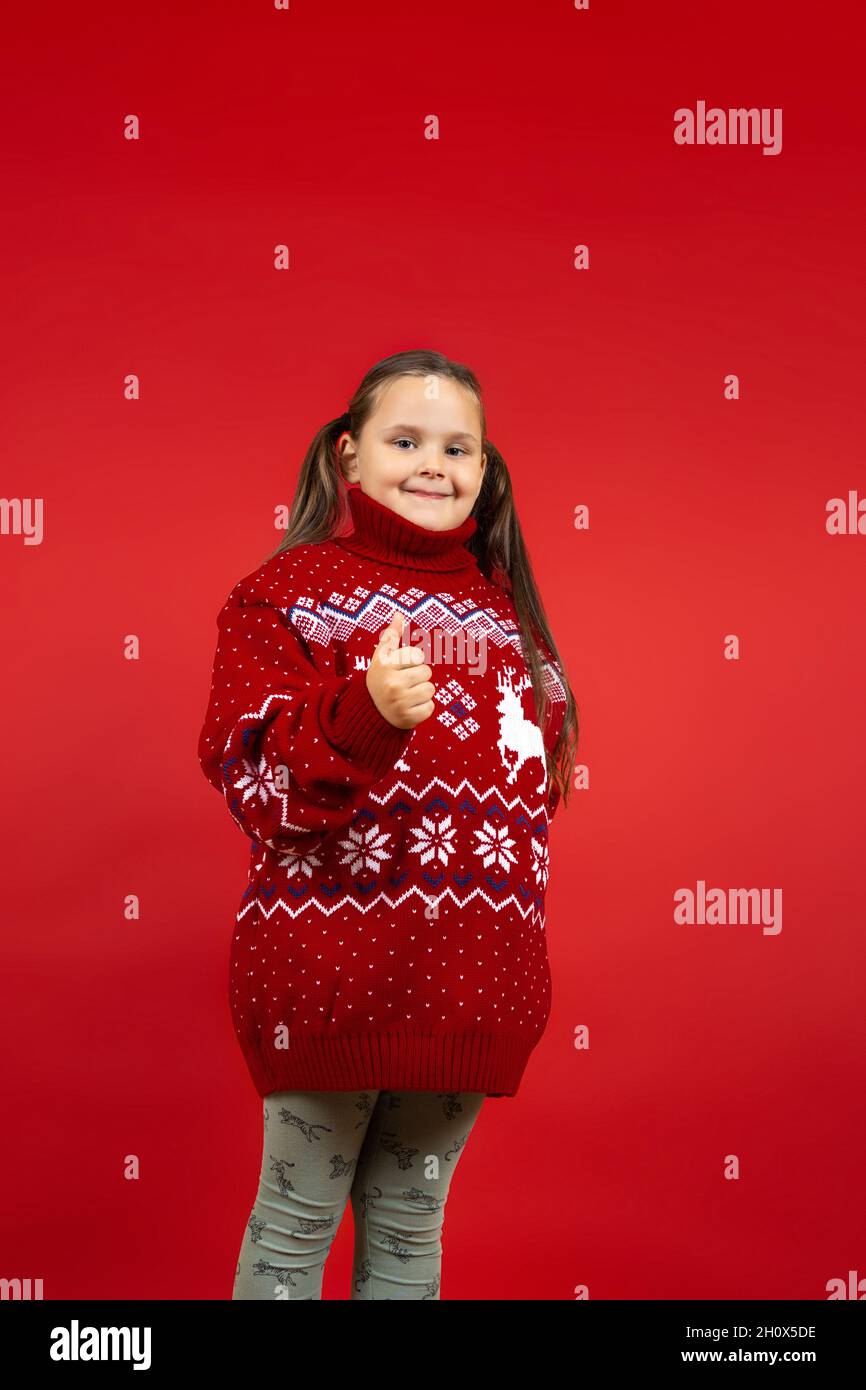 full-length portrait of smiling girl in red knitted Christmas sweater with reindeer giving a thumbs up, isolated on red background Stock Photo