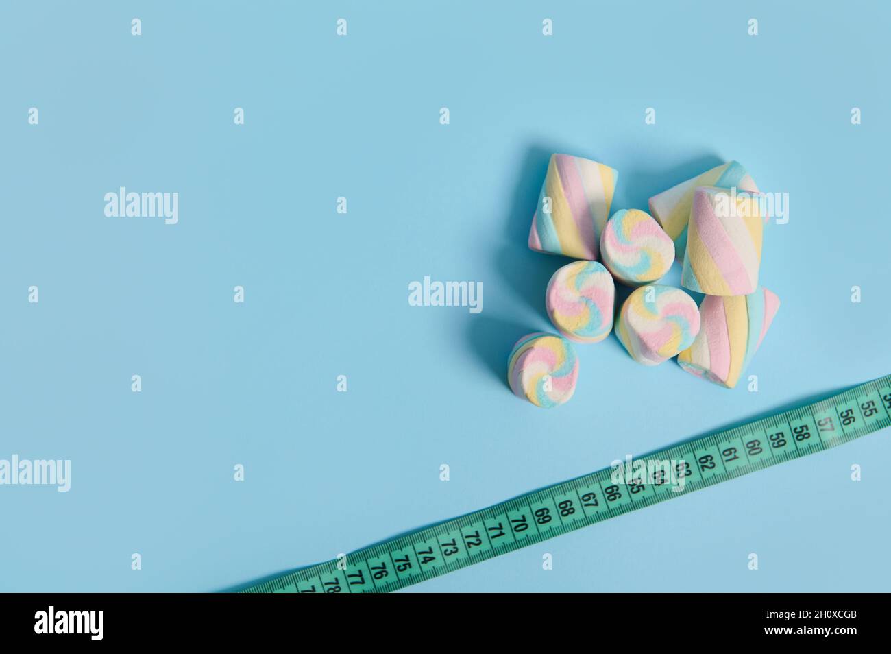 Top view of sweet colorful marshmallows and measuring tape on corner of blue background with copy space for medical ad. Link between overweight and un Stock Photo