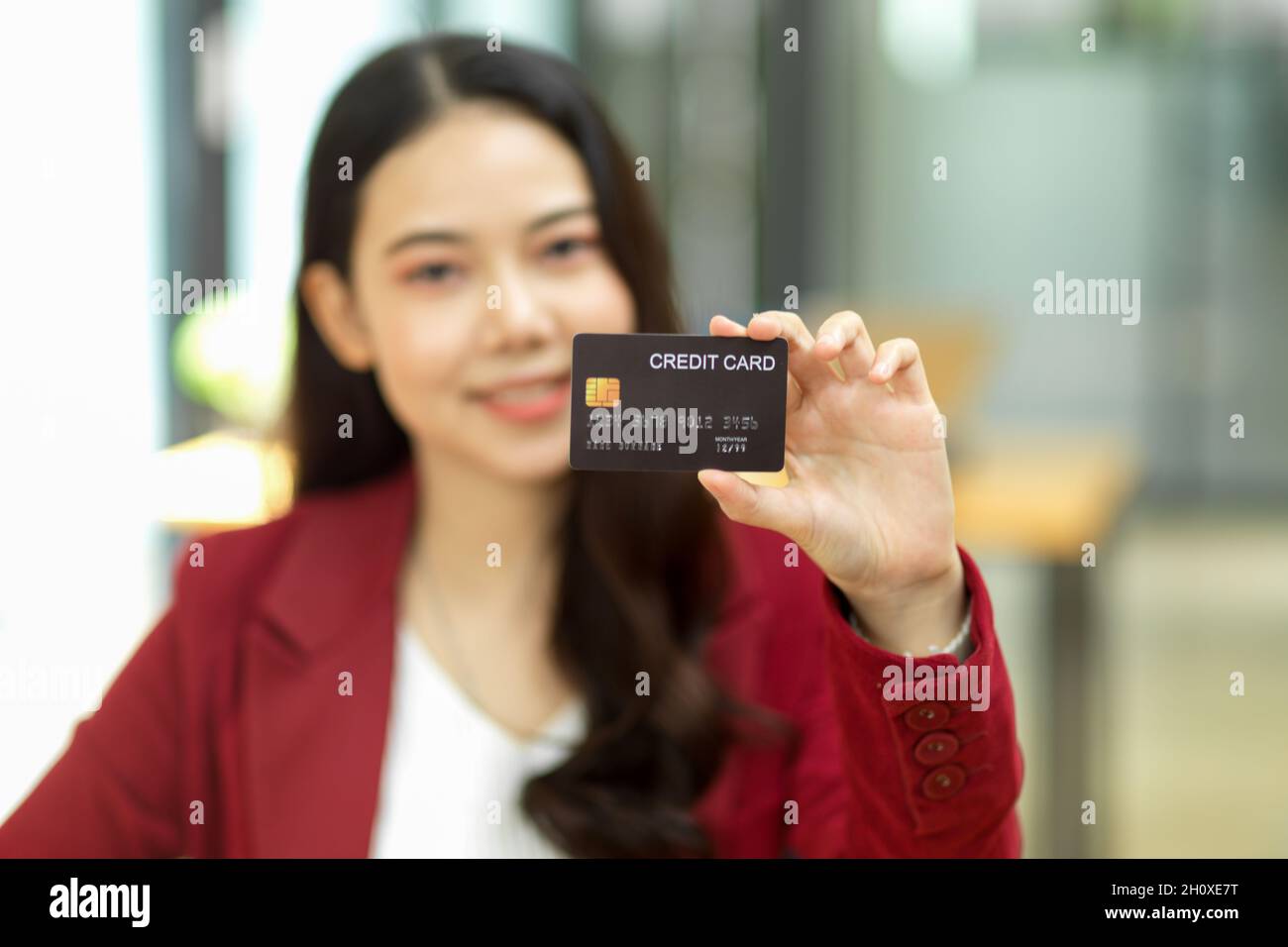 Beautiful asian female business worker in red suit with a credit card and blurred office in the background. cashless, online payment, internet banking Stock Photo