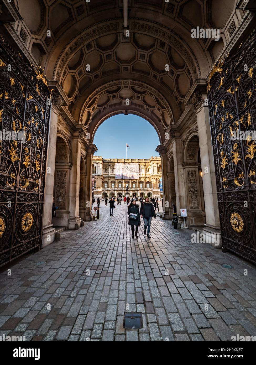 Royal Academy of Arts, London. A wide angle view through the entrance arch and gates of the RA with visitors leaving an exhibition of Anthony Gormley. Stock Photo