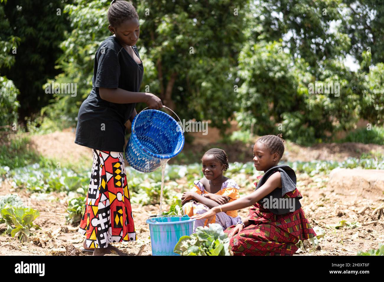 Troubled young black African women pouring water into a bucket at the village well with two little girls waiting for the container to be filled Stock Photo