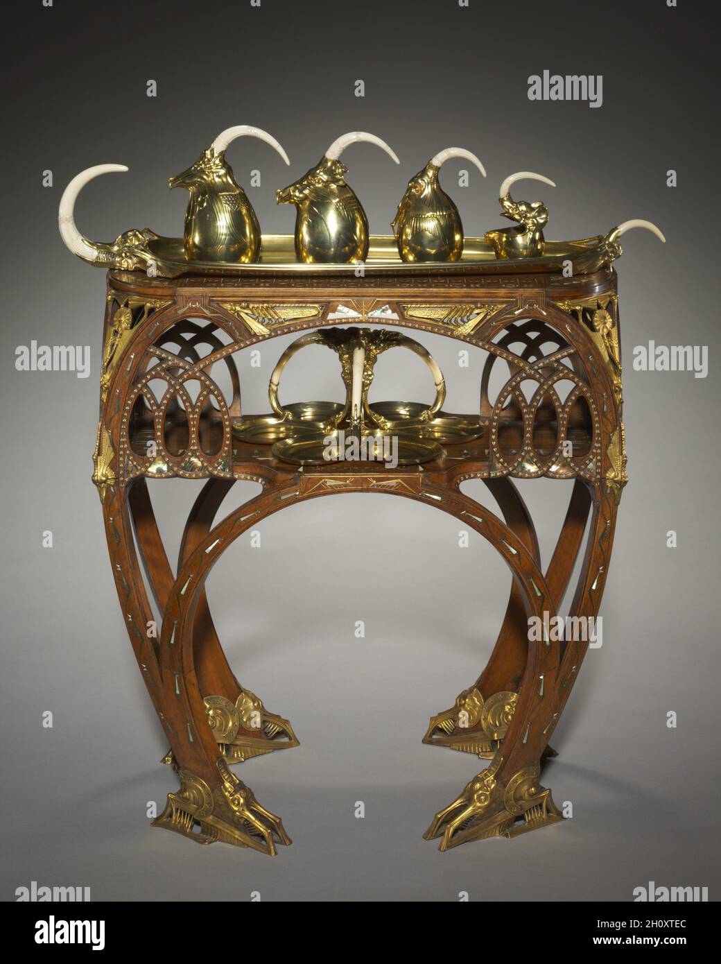 Tea Table, c. 1907. Carlo Bugatti (Italian, 1856-1940). Inlaid wood (mahogany?), cast and gilded bronze mounts, inlays of ivory or bone, metal, and mother-of-pearl (marine mussels or pearl oysters); overall: 71.5 x 67.1 x 41.3 cm (28 1/8 x 26 7/16 x 16 1/4 in.).  Luxurious materials might catch the eye first, but a closer look reveals the imaginary creatures that adorn the surface of this table. Featuring elaborate arches, a favorite motif that appeared throughout Bugatti's furniture, this fantastical imagery is a window into the designer's expressive imagination. Stock Photo