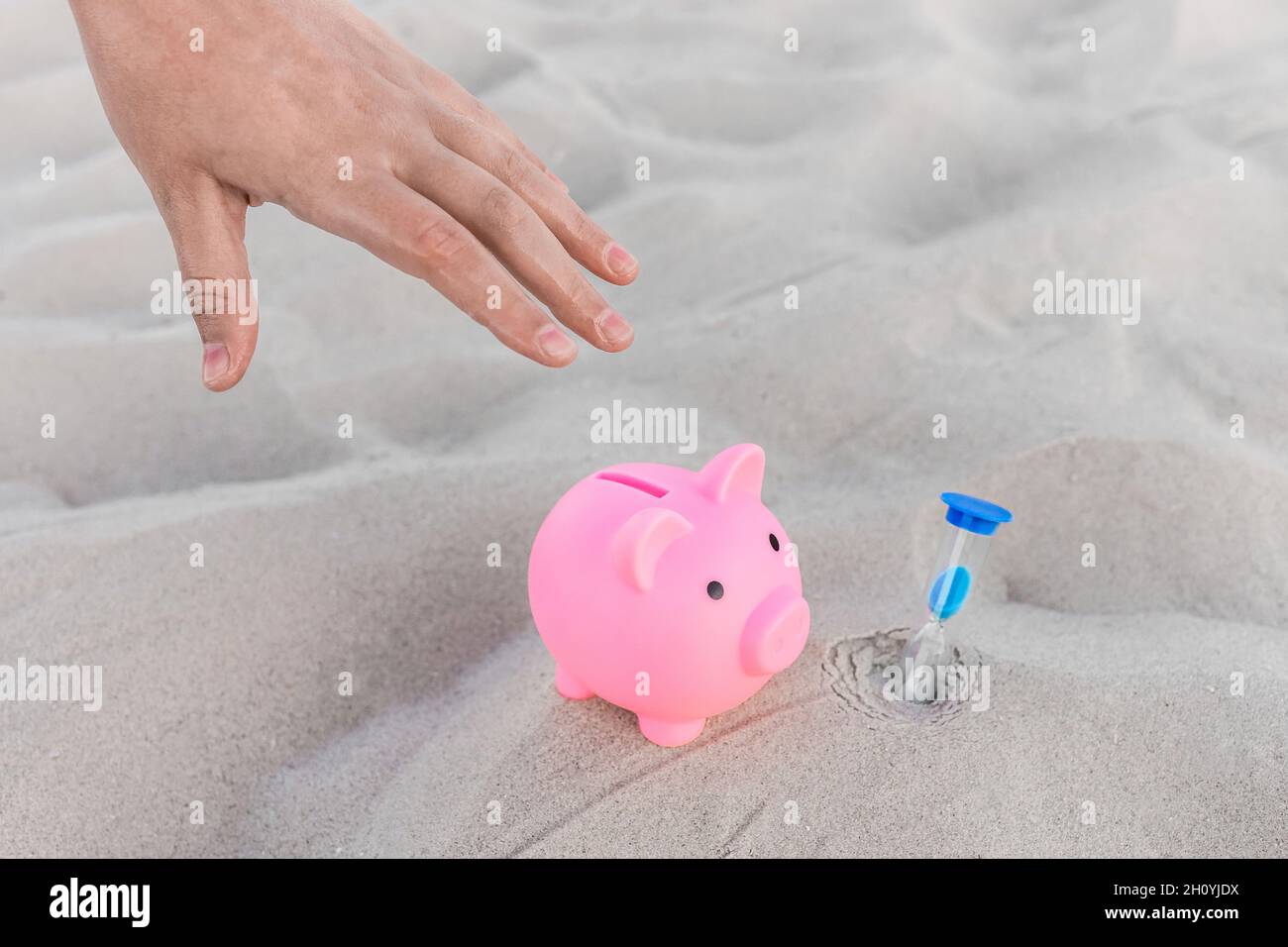 A human hand takes a piggy bank next to an hourglass against the background of white beach sand. Money, investments and fanance deposits for holidays Stock Photo
