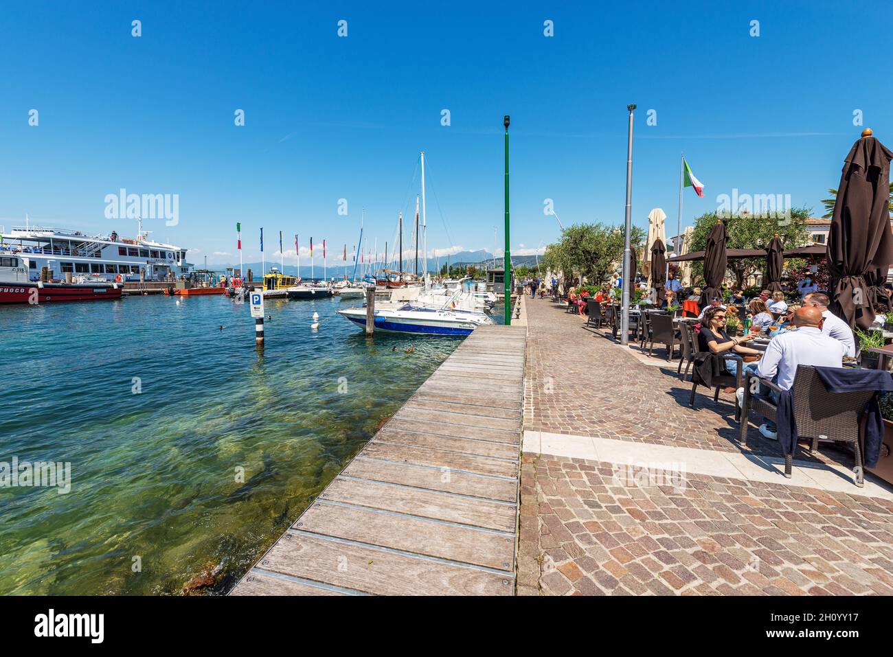 Port of the small village of Bardolino with many boats moored. Outdoors restaurants with many tourists. Lake Garda. Verona province, Veneto, Italy. Stock Photo