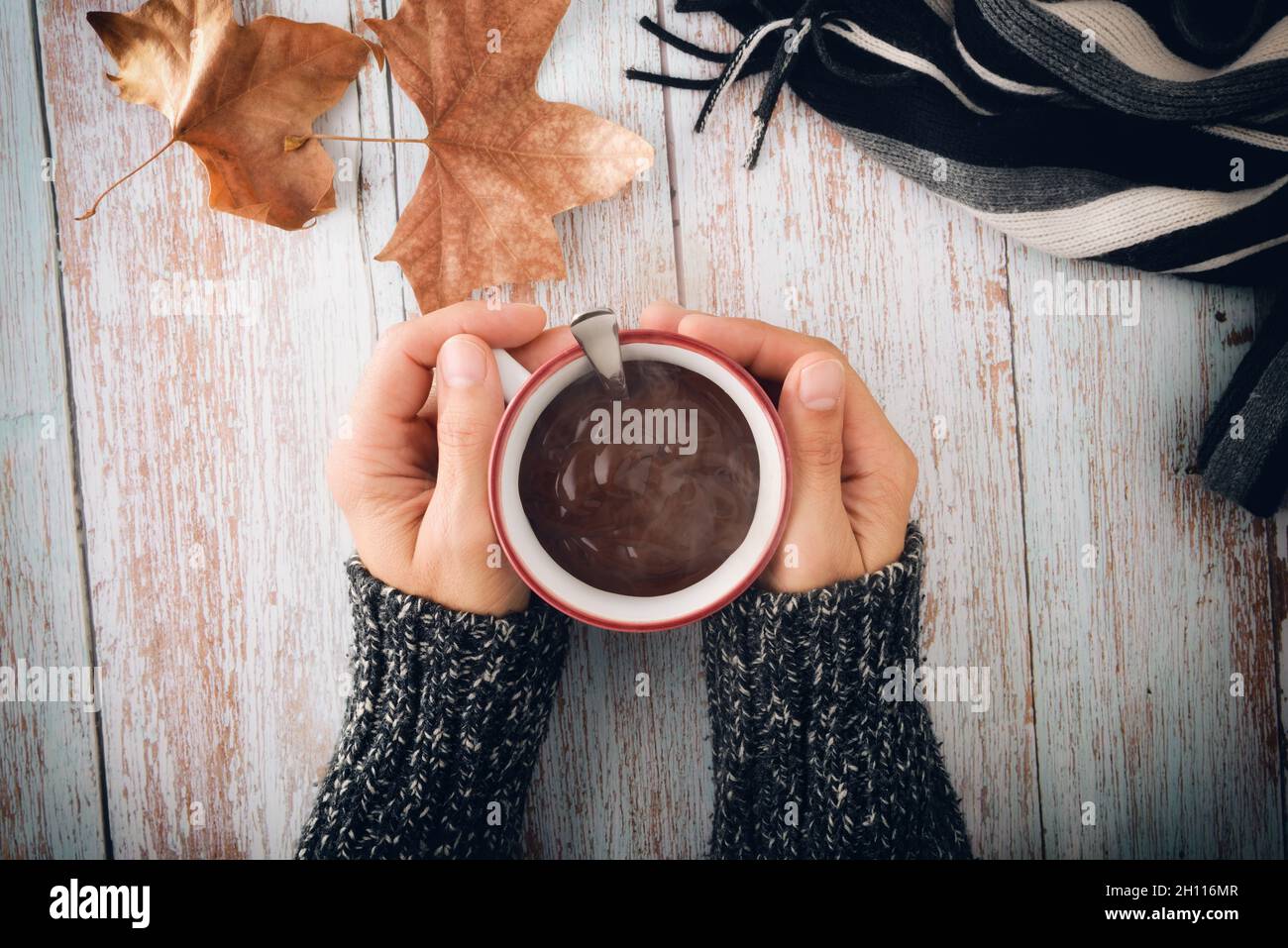 Cold hands in wool sweater catching cup of hot chocolate on wooden table with scarf and dry leaves in autumn. Top view Stock Photo