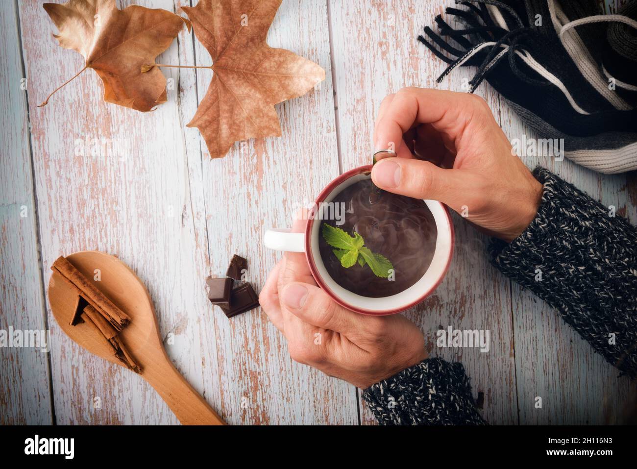 Cold hands with wool sweater stirring hot chocolate in a mug on wooden table with chocolate and wooden spoon with cinnamon and scarf and dry leaves in Stock Photo