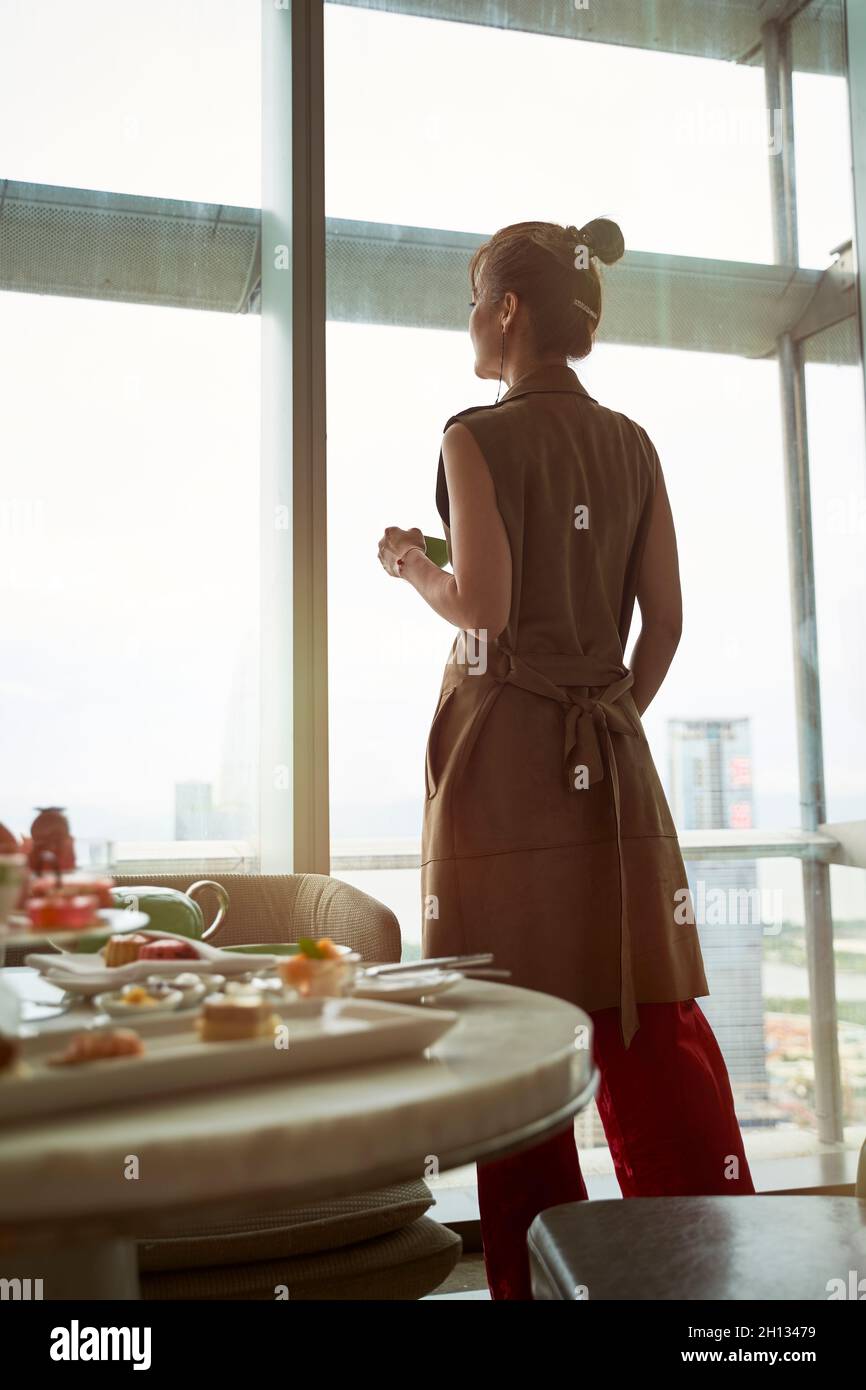 mature asian woman looking at view through window of hotel room with breakfast on table Stock Photo