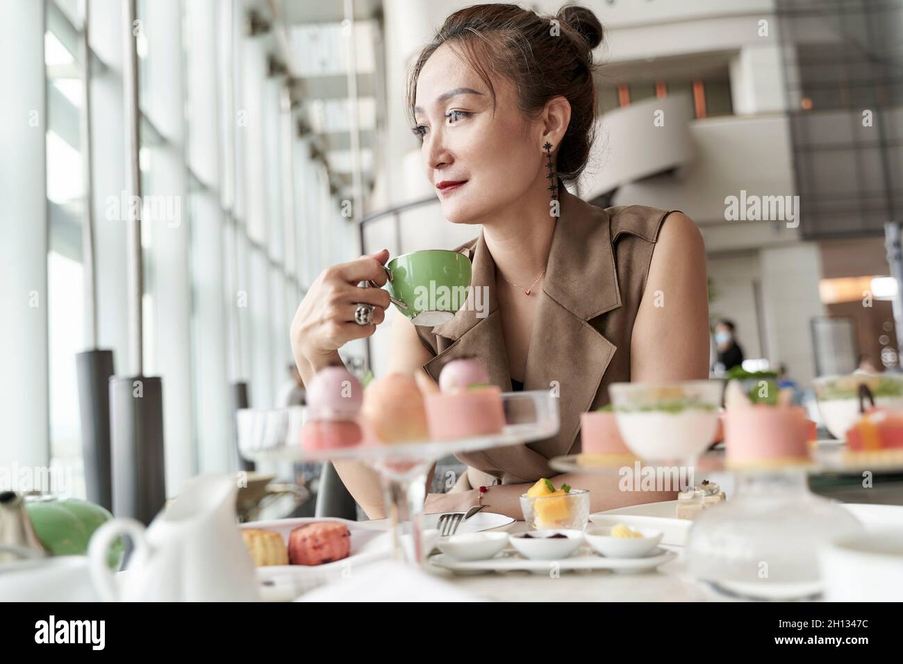 mature asian woman sitting at table enjoying afternoon tea in hotel lobby coffee shop Stock Photo