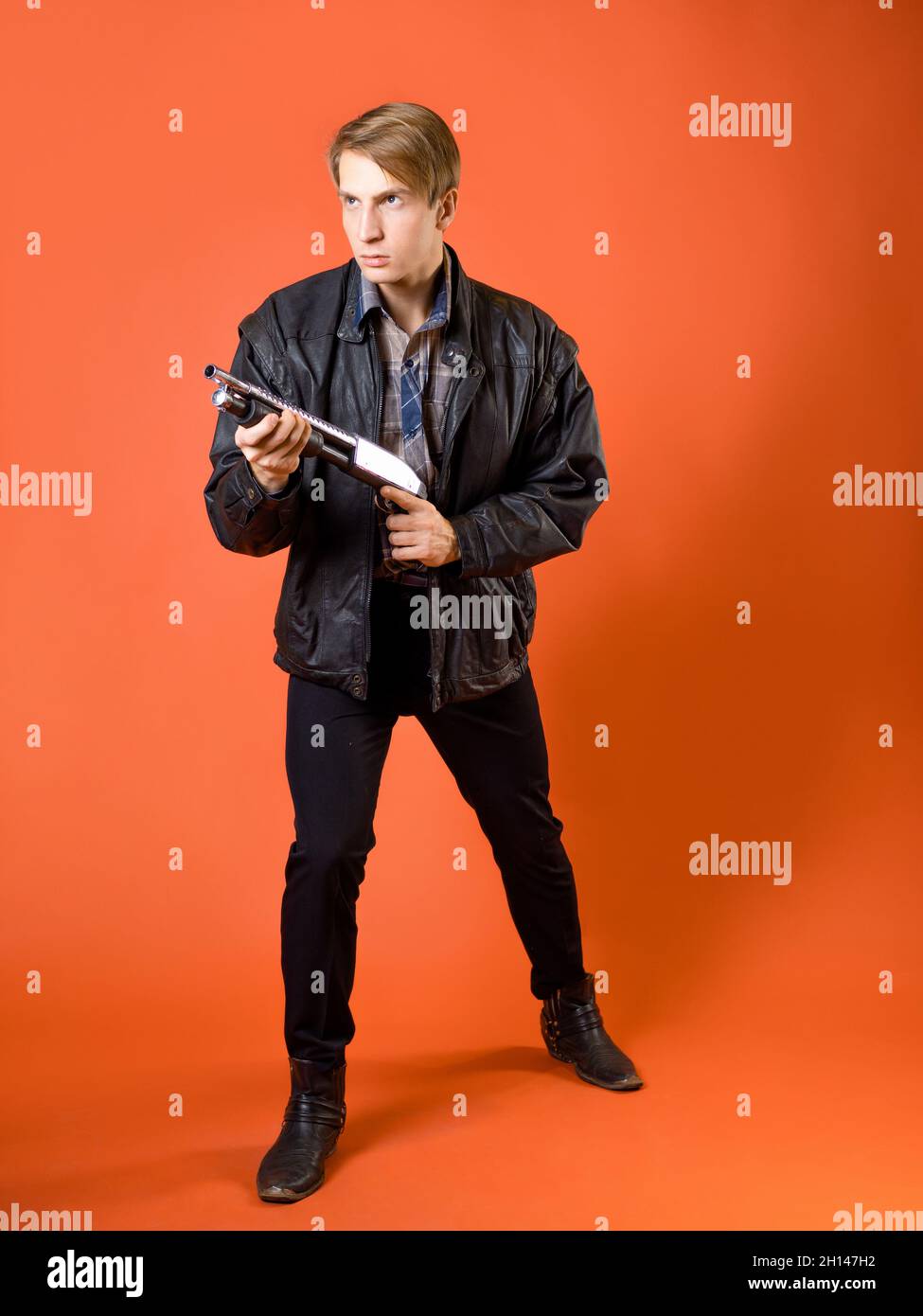 a cool hunter of evil spirits, a guy in a casual shirt and a leather jacket with a shotgun, ready to attack, a defensive stance, a studio photo on an Stock Photo