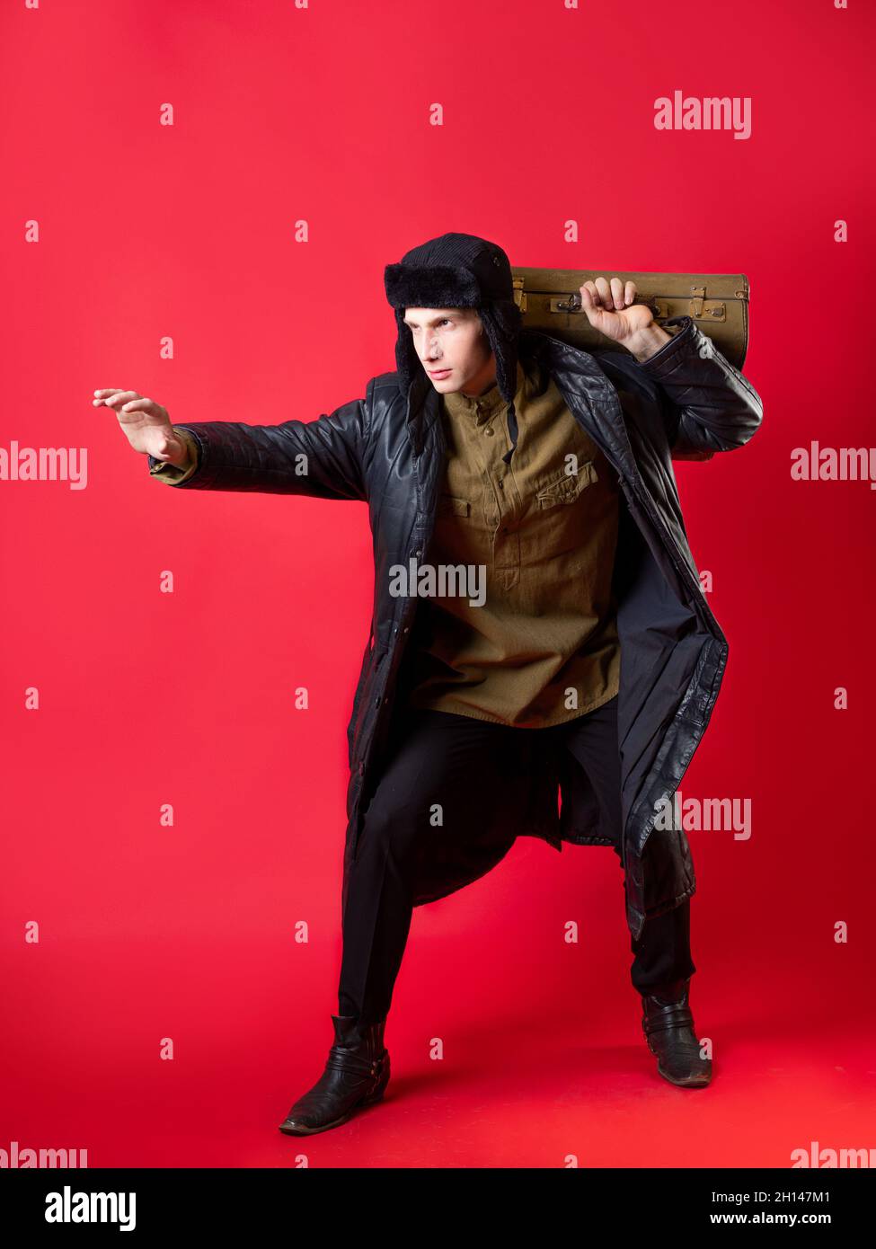 A man in old-fashioned clothes and a hat with earflaps he drags his big suitcase on himself, Russian post-war style. Portrait on a red background Stock Photo