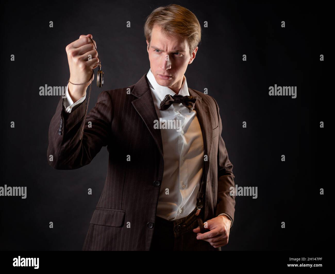 An intelligent gentleman in the Victorian style. Vintage retro suit, young attractive man in a vest and bow tie, he holds a pocket watch on a chain in Stock Photo