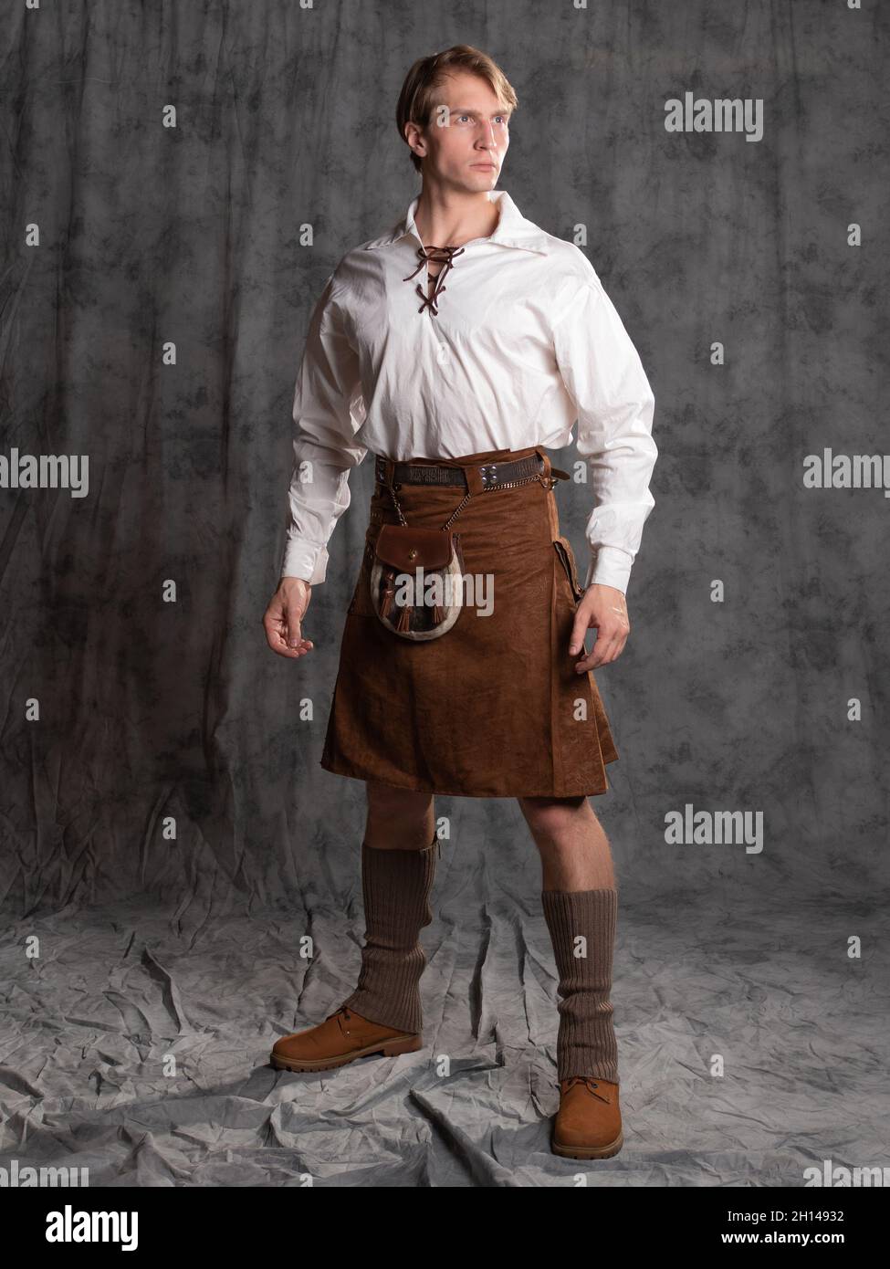 A young man in a leather kilt and a white lace-up blouse. A Scottish, Photo in the studio on a gray background Stock Photo