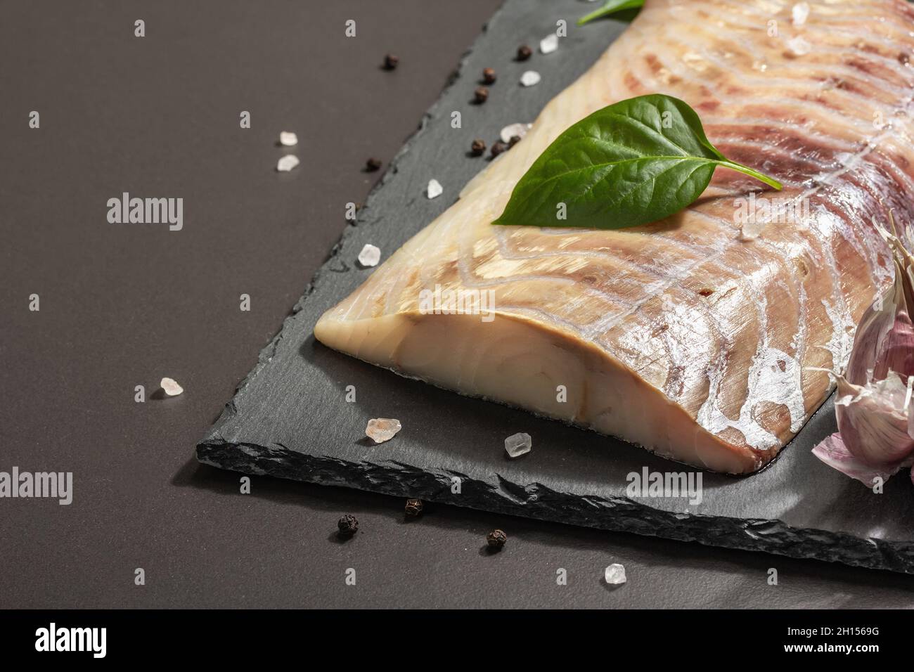 Raw pollock (Pollachius virens) fillet. Fresh fish for healthy food lifestyle. Garlic, sea salt, peppercorn, greens. Black stone concrete background, Stock Photo