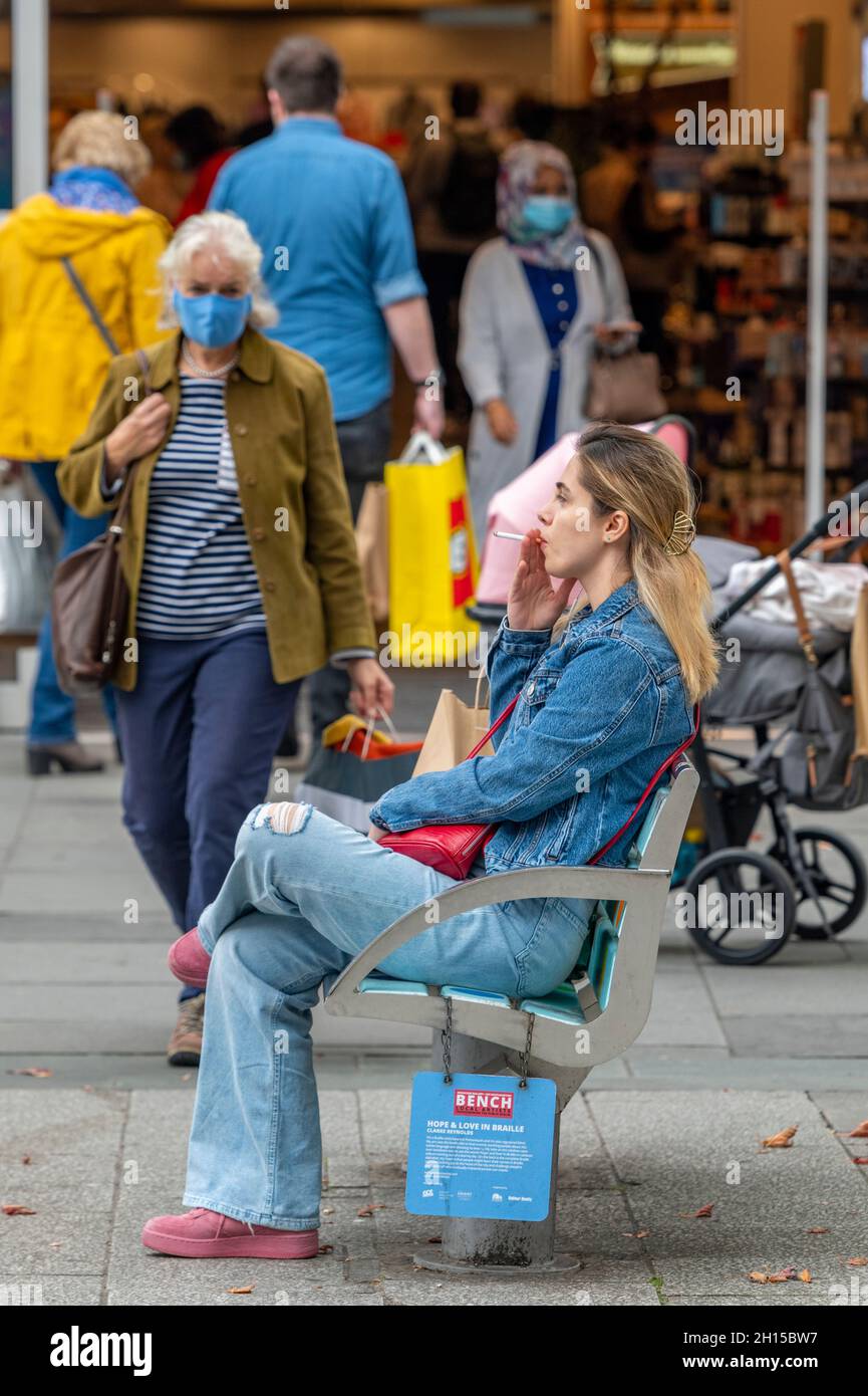 young woman sitting on a wooden bench in a busy town centre smoking a cigarette. young female smoking, smoker sitting on bench, young female smoker. Stock Photo