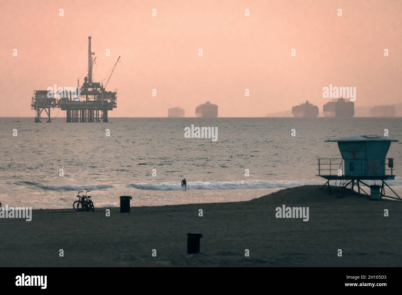 A lone surfer comes in on a wave at Huntingdon beach, oil rig and c ontainer ships dot the horizon at sunset. Stock Photo