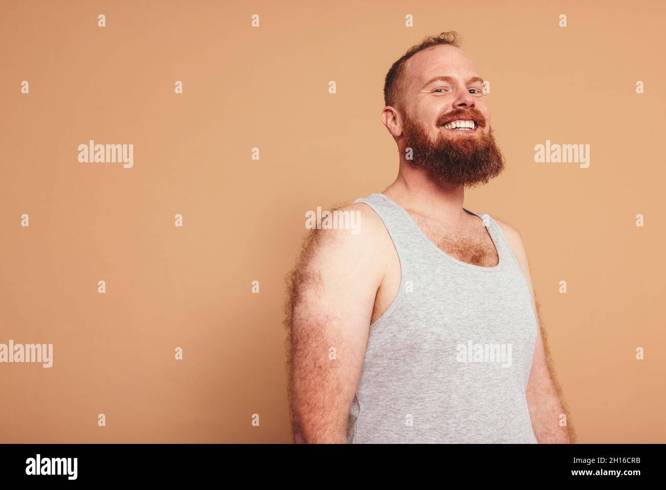 Man with a ginger beard smiling at the camera cheerfully. Body positive young man standing against a studio background. Self-assured young man feeling Stock Photo