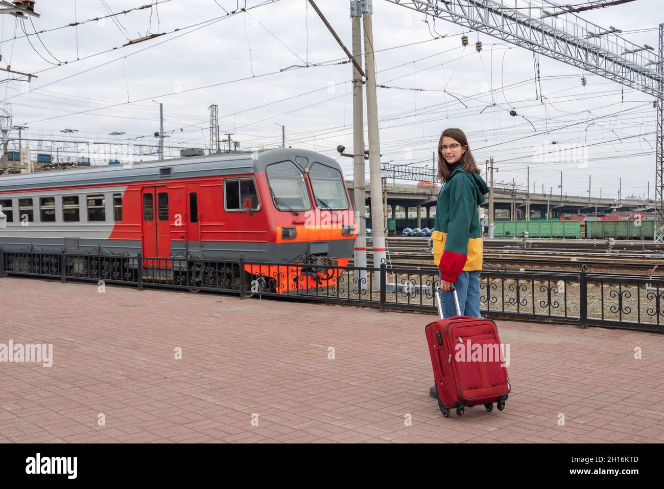 Caucasian girl student in bright clothes with a suitcase going to the electric train at the station. Girl with a suitcase at the train station. Depart Stock Photo