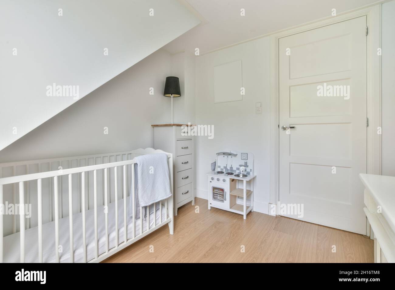 White crib with towel placed in light stylish bedroom with lamp on chest of drawers and toy near closed door Stock Photo