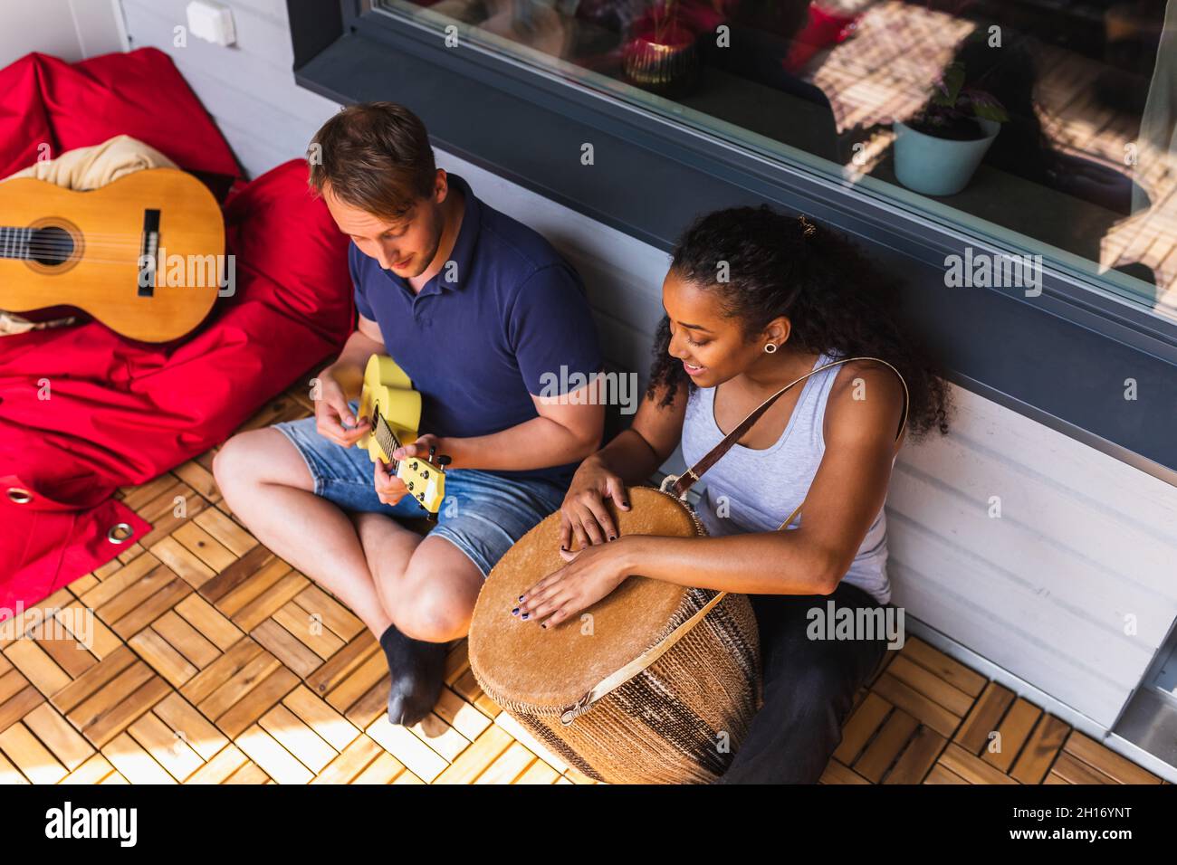From above of cheerful young diverse couple resting and playing ukulele and djembe sitting on