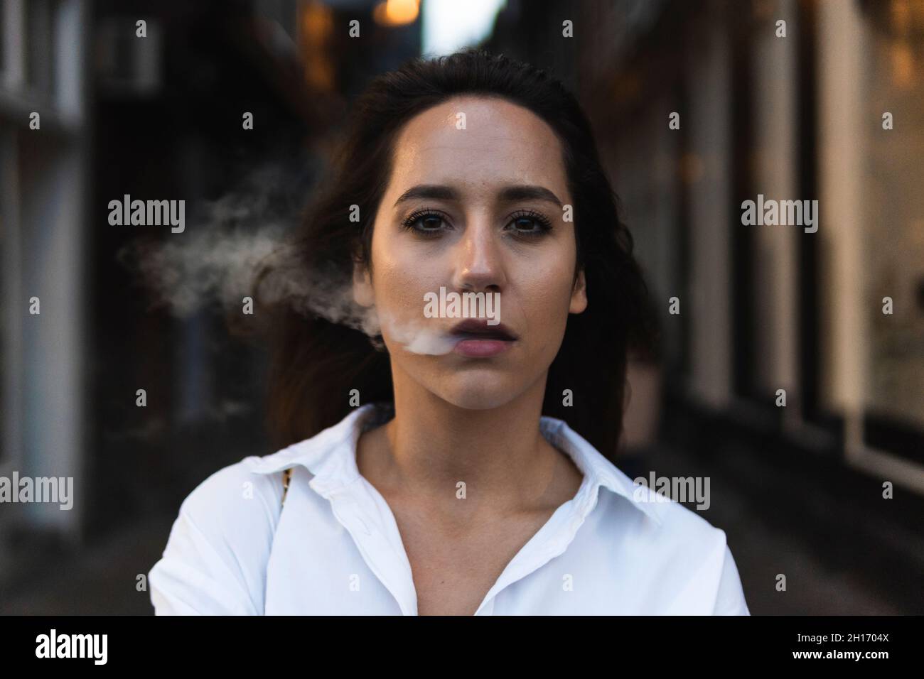 Confident female in white shirt exhaling smoke while standing on street and looking at camera Stock Photo