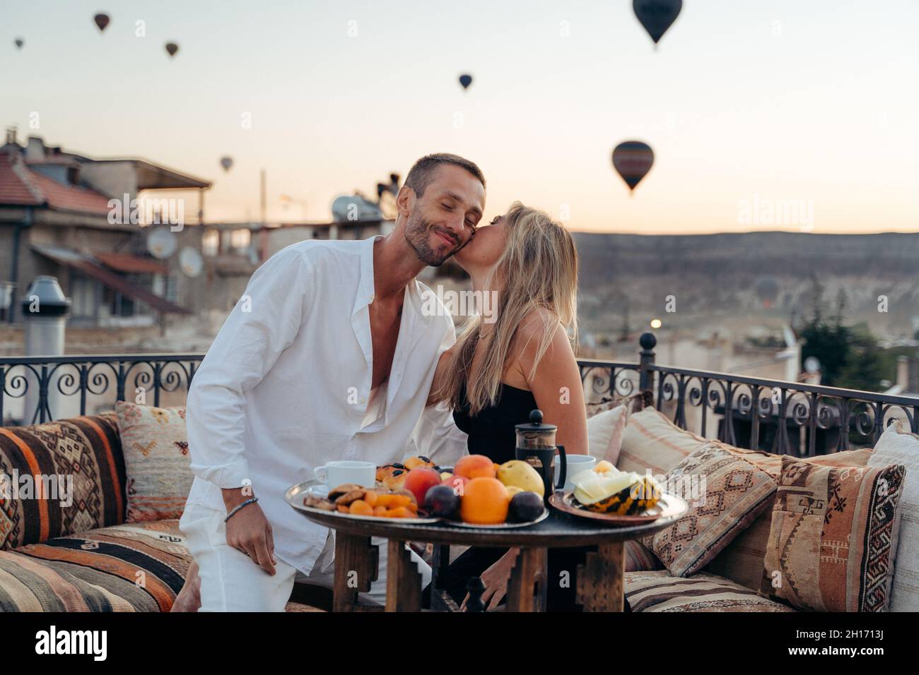 Woman kissing soulmate while sitting in cafe on rooftop terrace and enjoying summer evening on background of hot air balloons Stock Photo