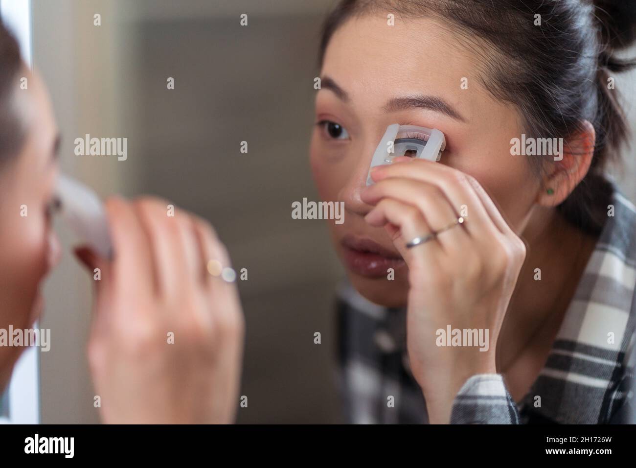 Charming concentrated Asian female using curler on eyelashes while doing makeup in front of mirror at home Stock Photo