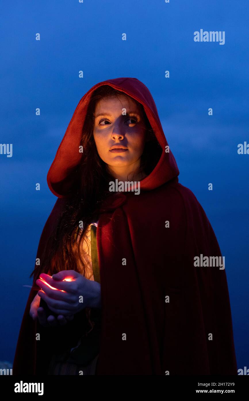 Serious female in Victorian styled outfit with red cape looking away while standing with glowing candle in hands in dark nature Stock Photo