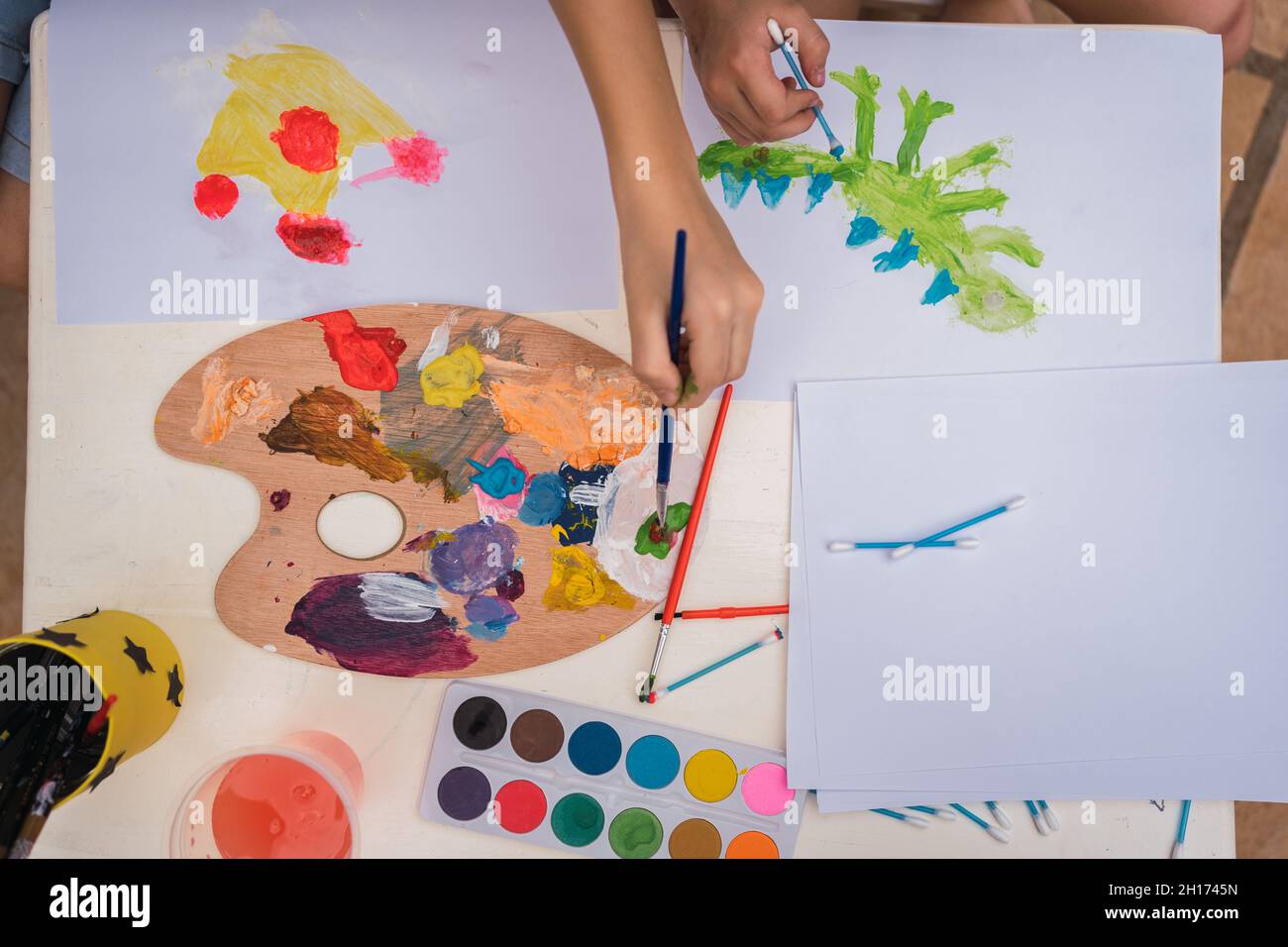 From above of crop anonymous children painting on white papers with ...