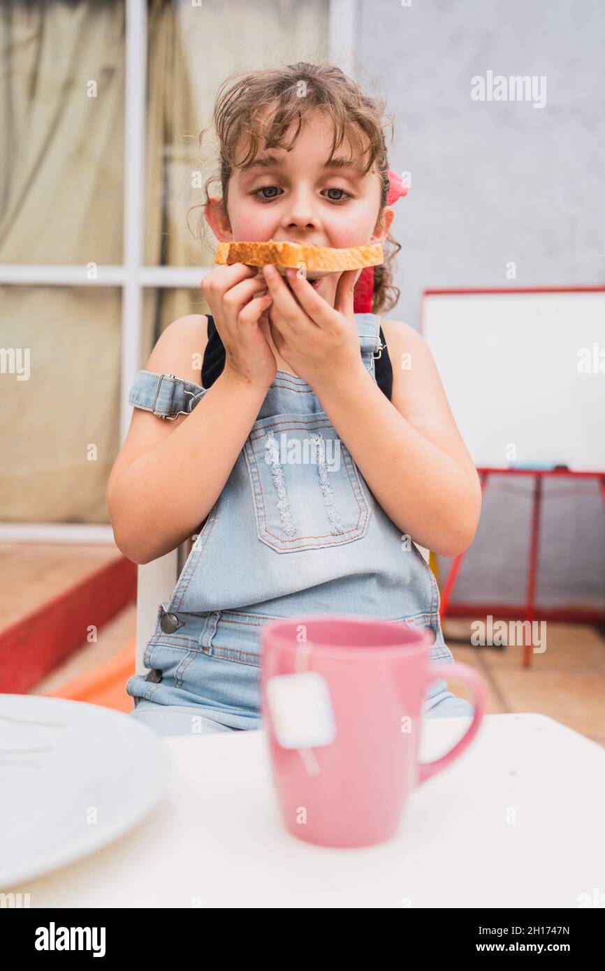 Cute girl in denim overall looking down while eating fresh bread with sweet  jam in light room at home Stock Photo - Alamy
