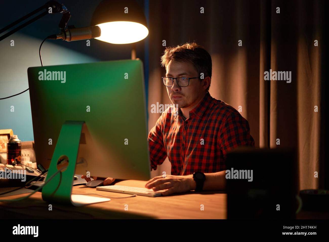 Concentrated male in checkered shirt and eyeglasses working on computer sitting at table with lamp and microphone during recording podcast Stock Photo