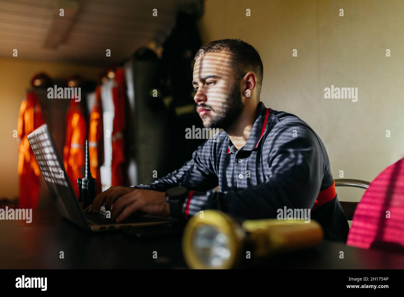 Concentrated young bearded ethnic guy wearing shirt sitting at table with laptop and transmitter looking at screen with yellow flashlight in foregroun Stock Photo