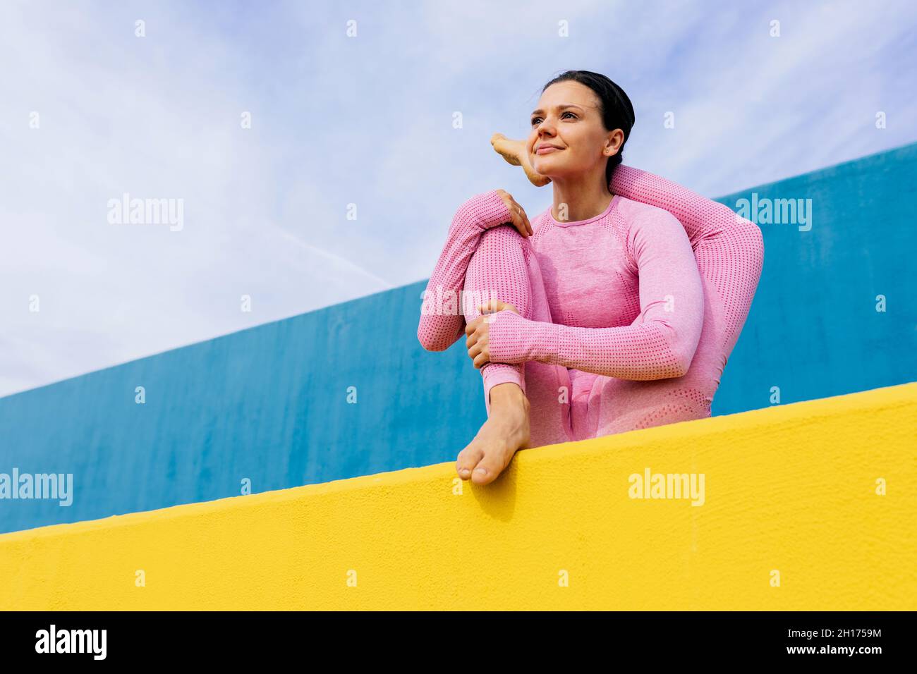 Young pensive female doing variation of Baby Cradle pose while meditating in yoga asana on blue and yellow background Stock Photo