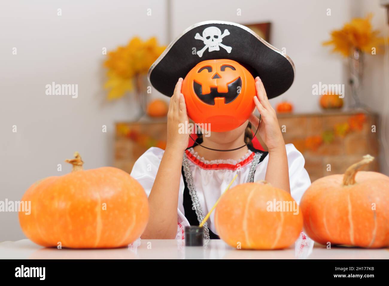 Halloween. A funny little girl in a pirate costume hides her head behind a bucket of pumpkin with a lantern. The child sees at home at the table again Stock Photo
