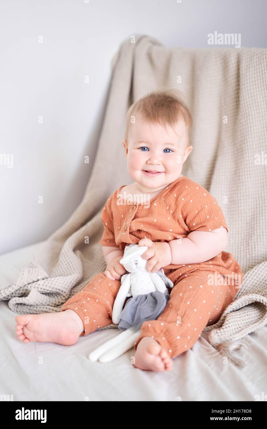Portrait of a cute 9 months old baby girl seating on a bed. playing with a bunny toy Stock Photo
