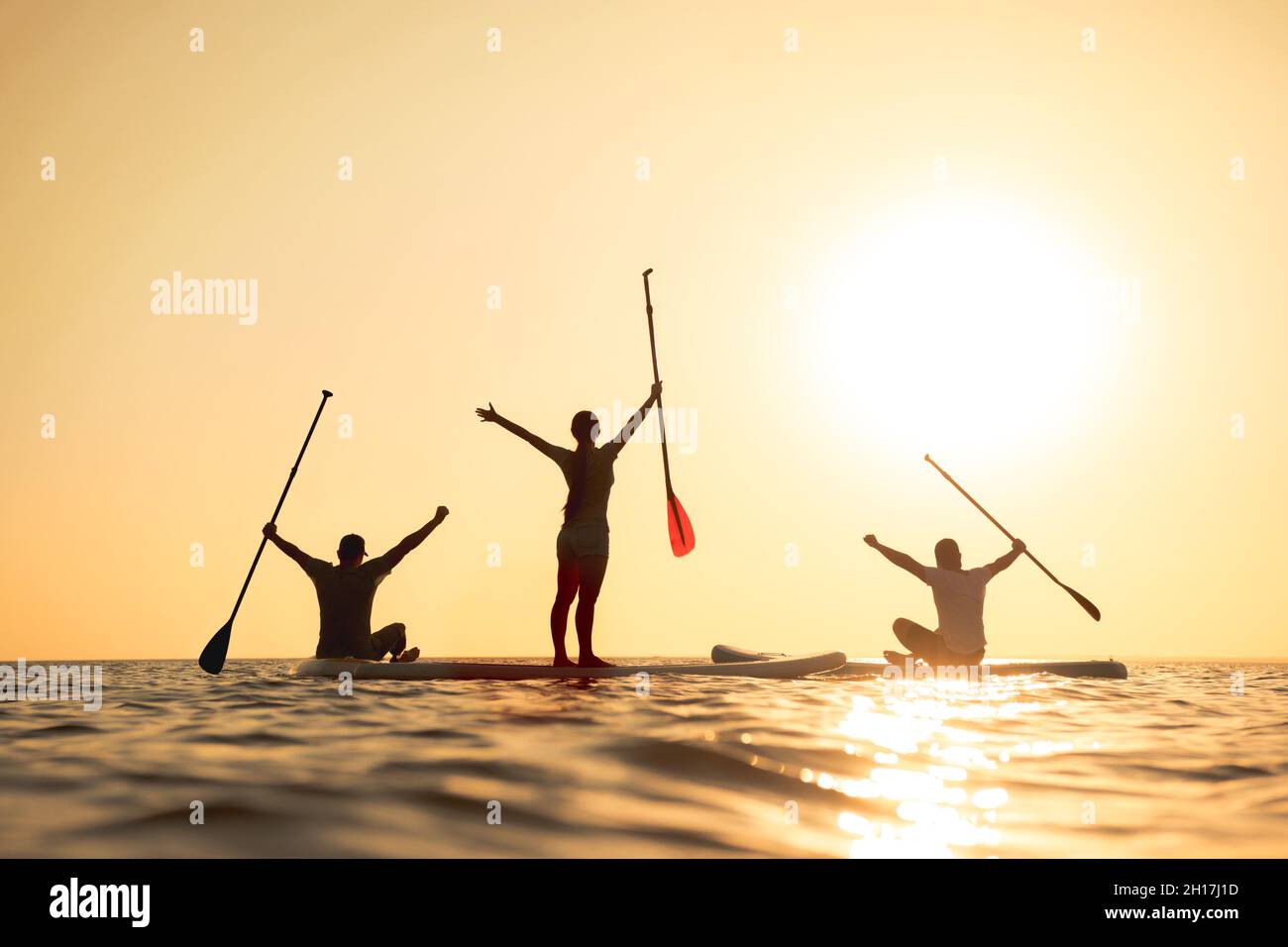 Happy surfers stands on sup boards with raised arms and looks at sunset Stock Photo