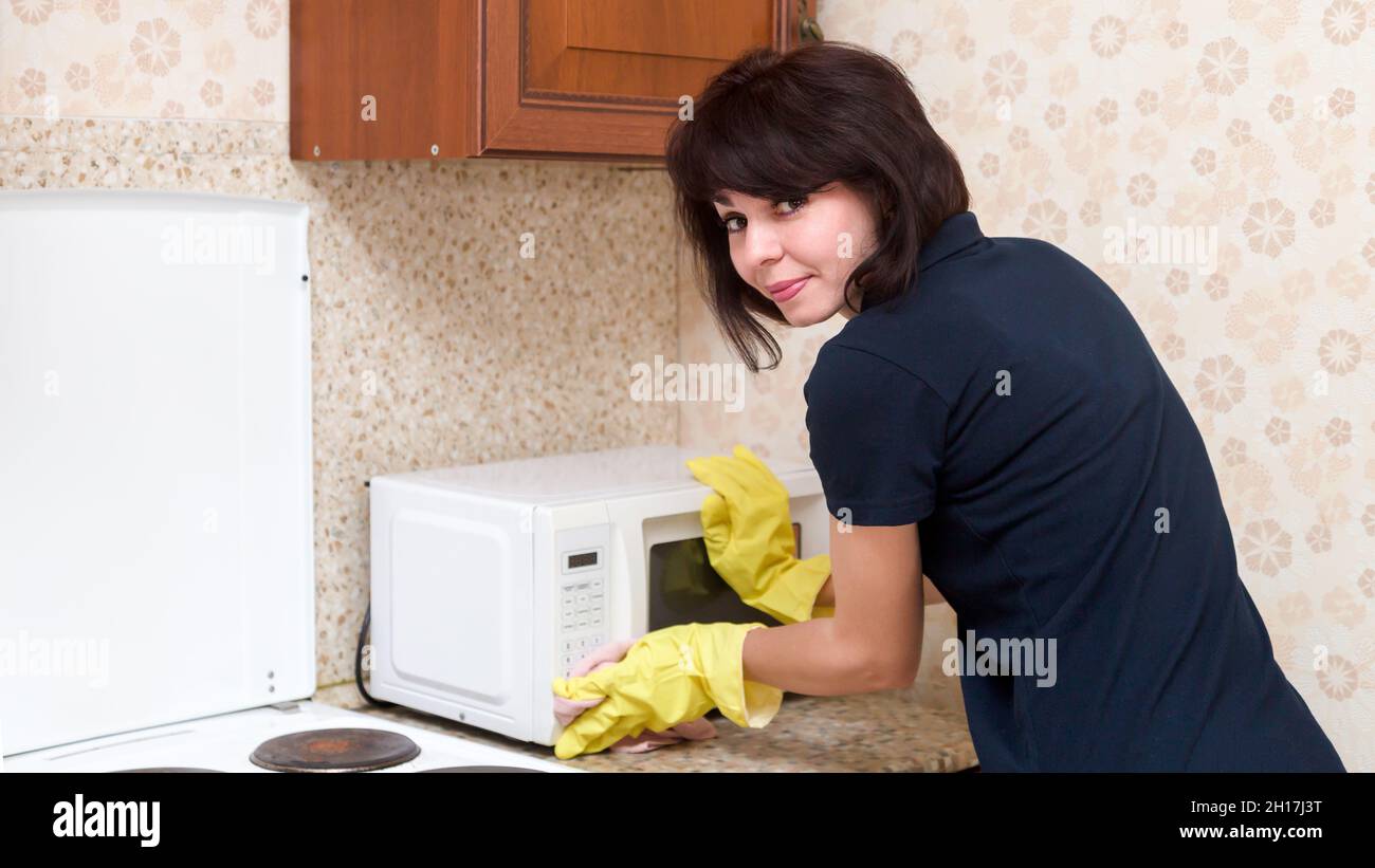 Brunette housewife in yellow rubber gloves washes kitchen microwave oven. Stock Photo