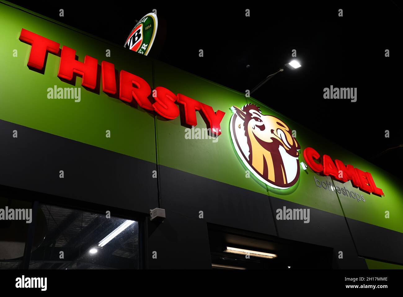 The illuminated sign of a Thirsty Camel Bottleshop at night, with a partially obscured Victoria Bitter sign above Stock Photo